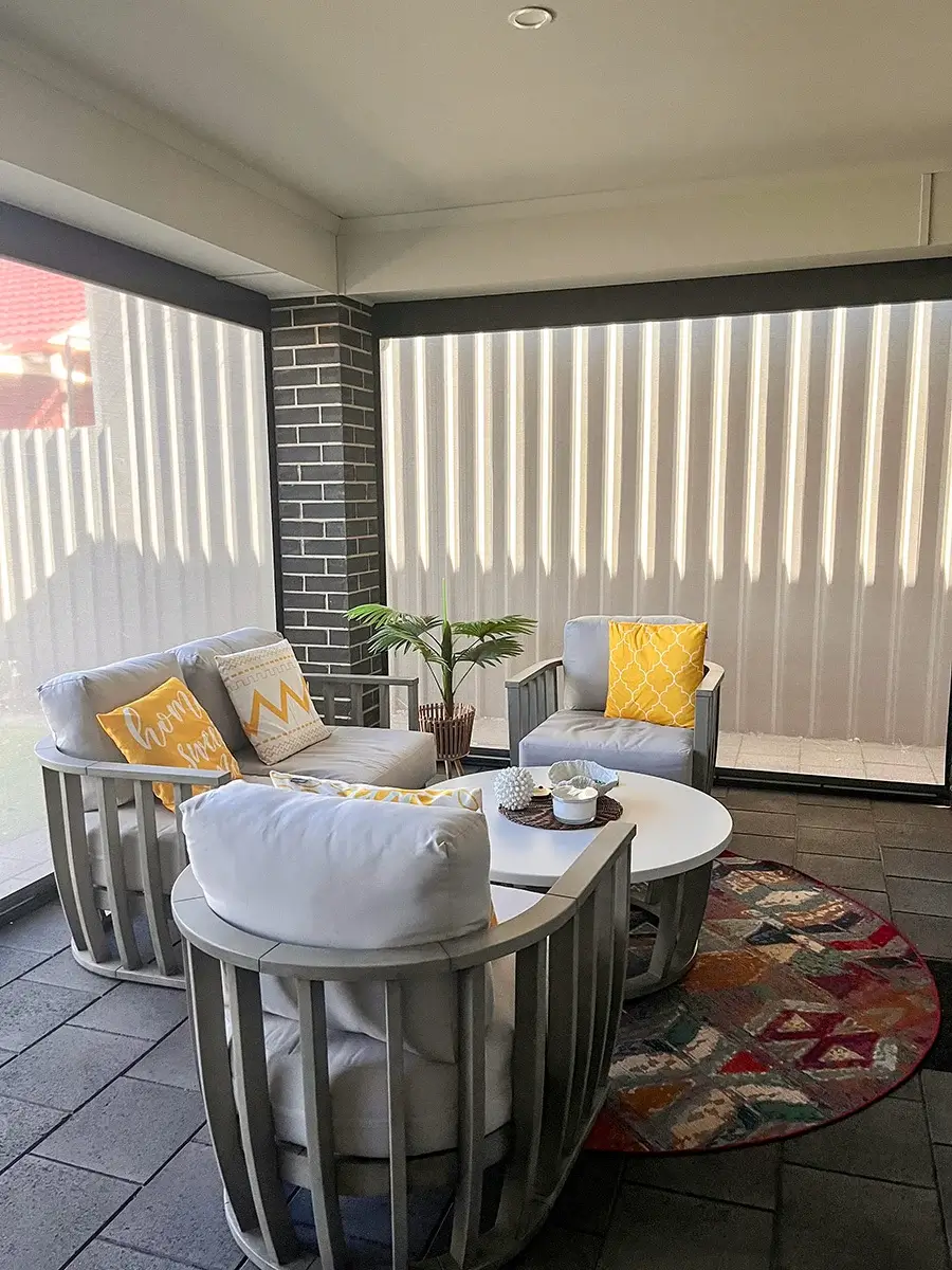 Modern outdoor seating area with Motorised Outdoor Blinds, featuring comfortable chairs with yellow and white cushions, a round table, and a colourful rug on a tiled floor.