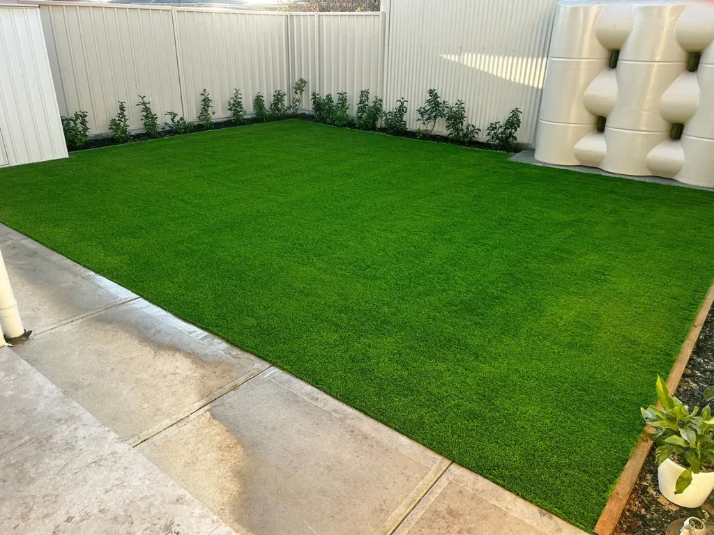 A neatly landscaped backyard featuring a lush artificial lawn. The yard is surrounded by a clean white fence with a row of young shrubs planted along the edges. A modern rainwater tank is positioned in the corner, and the ground is paved with concrete tiles, contributing to the well-organised and low-maintenance landscaping design.