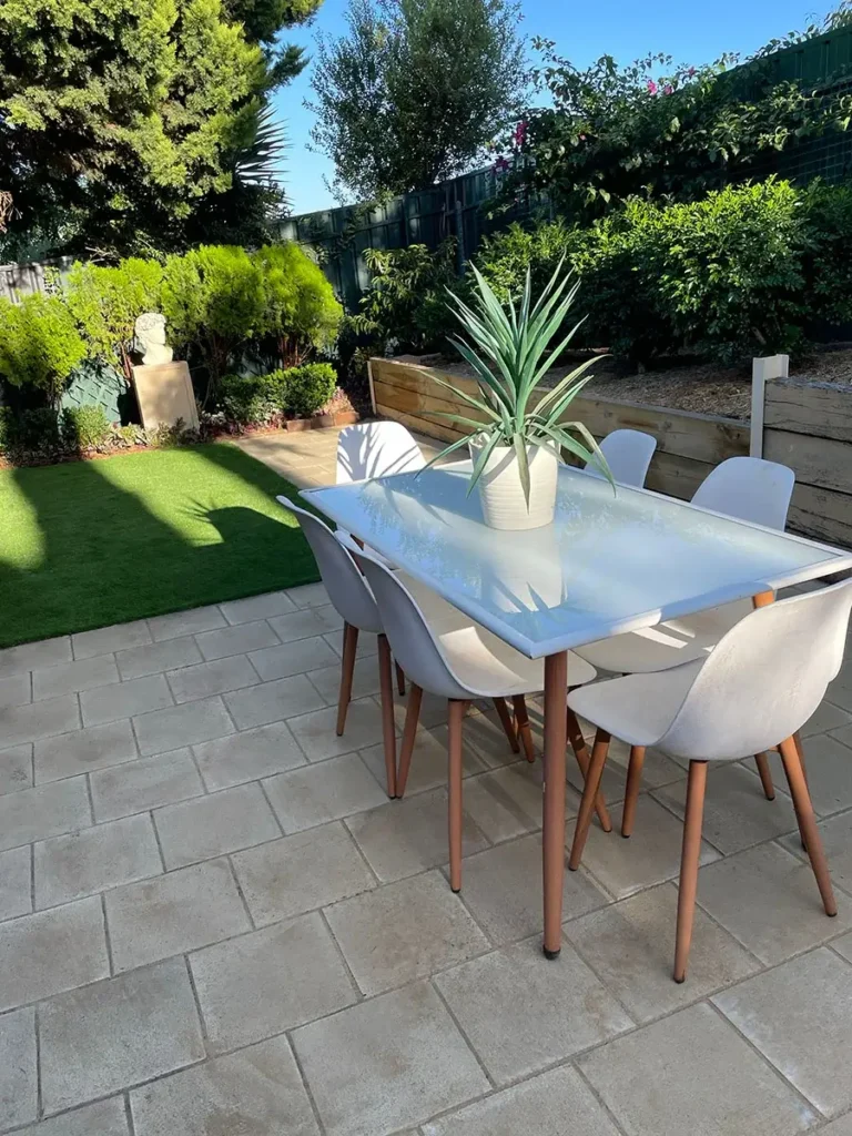 a backyard with a well-manicured lawn and a modern outdoor dining area. The dining set includes a glass-top table and white chairs with wooden legs, with a potted plant as a centrepiece. The surrounding garden features lush green shrubs, trees, and a raised wooden planter bed, creating a serene and inviting atmosphere.
