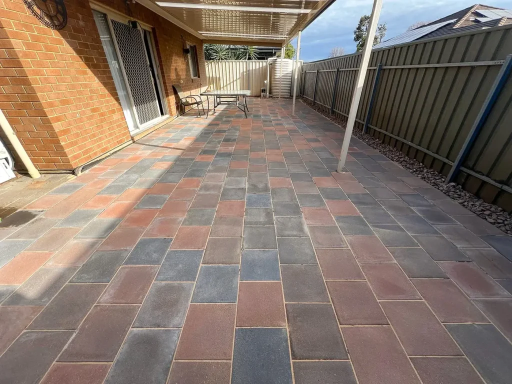 a backyard patio with a neatly laid brick paver floor in a pattern of alternating gray and red bricks. The patio is adjacent to a brick house with a sliding door and a covered roof. The area is enclosed by a green metal fence, and there is outdoor furniture including a table and chairs.