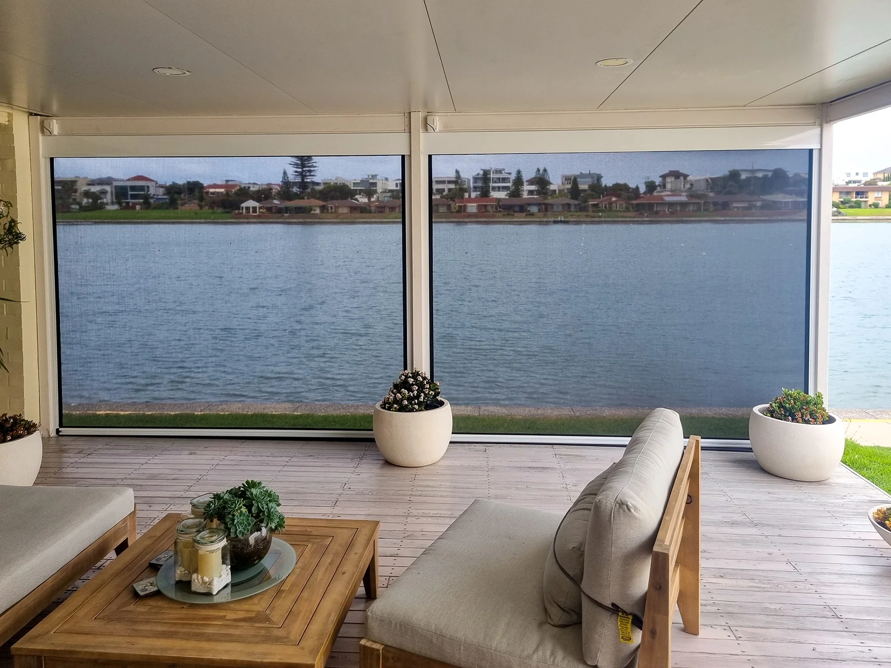 Cosy patio area with outdoor blinds, overlooking a serene waterfront, featuring comfortable seating, a wooden coffee table, and potted plants.