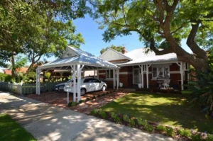 Charming brick house with a front yard and a white timber carport over the driveway, providing shade for parked cars, surrounded by lush trees and a picket fence.