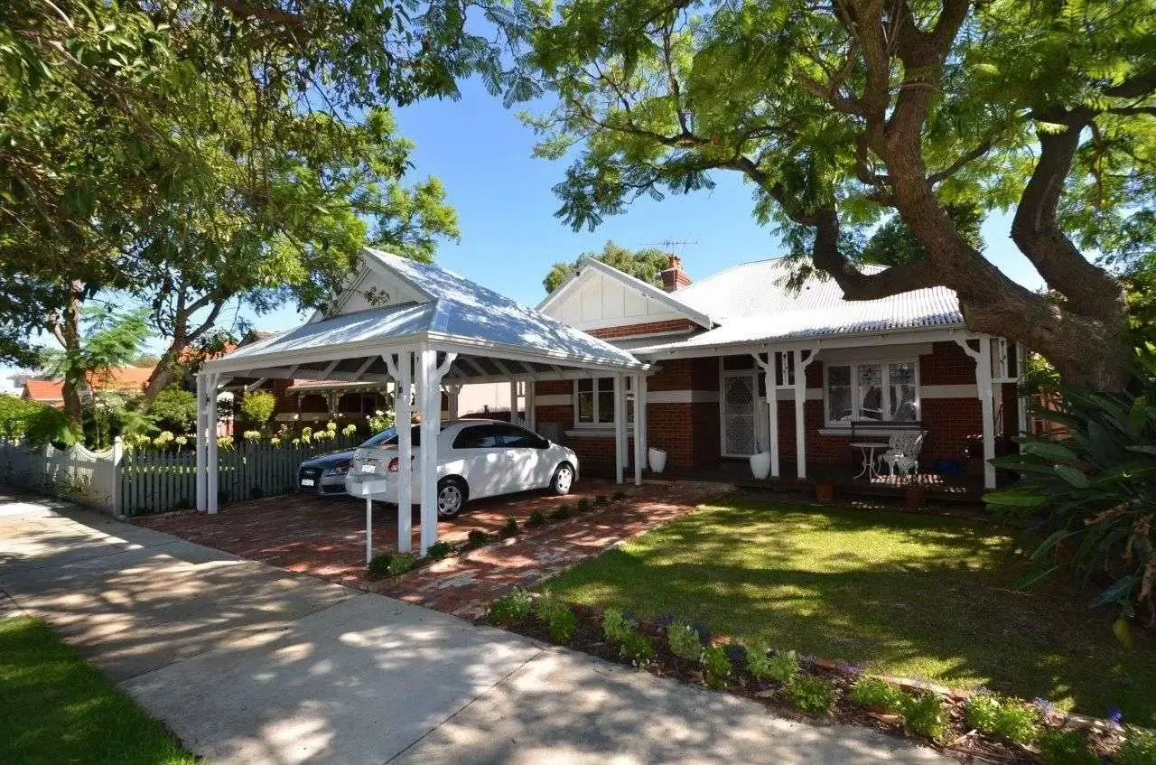 Charming brick house with a front yard and a white timber carport over the driveway, providing shade for parked cars, surrounded by lush trees and a picket fence.