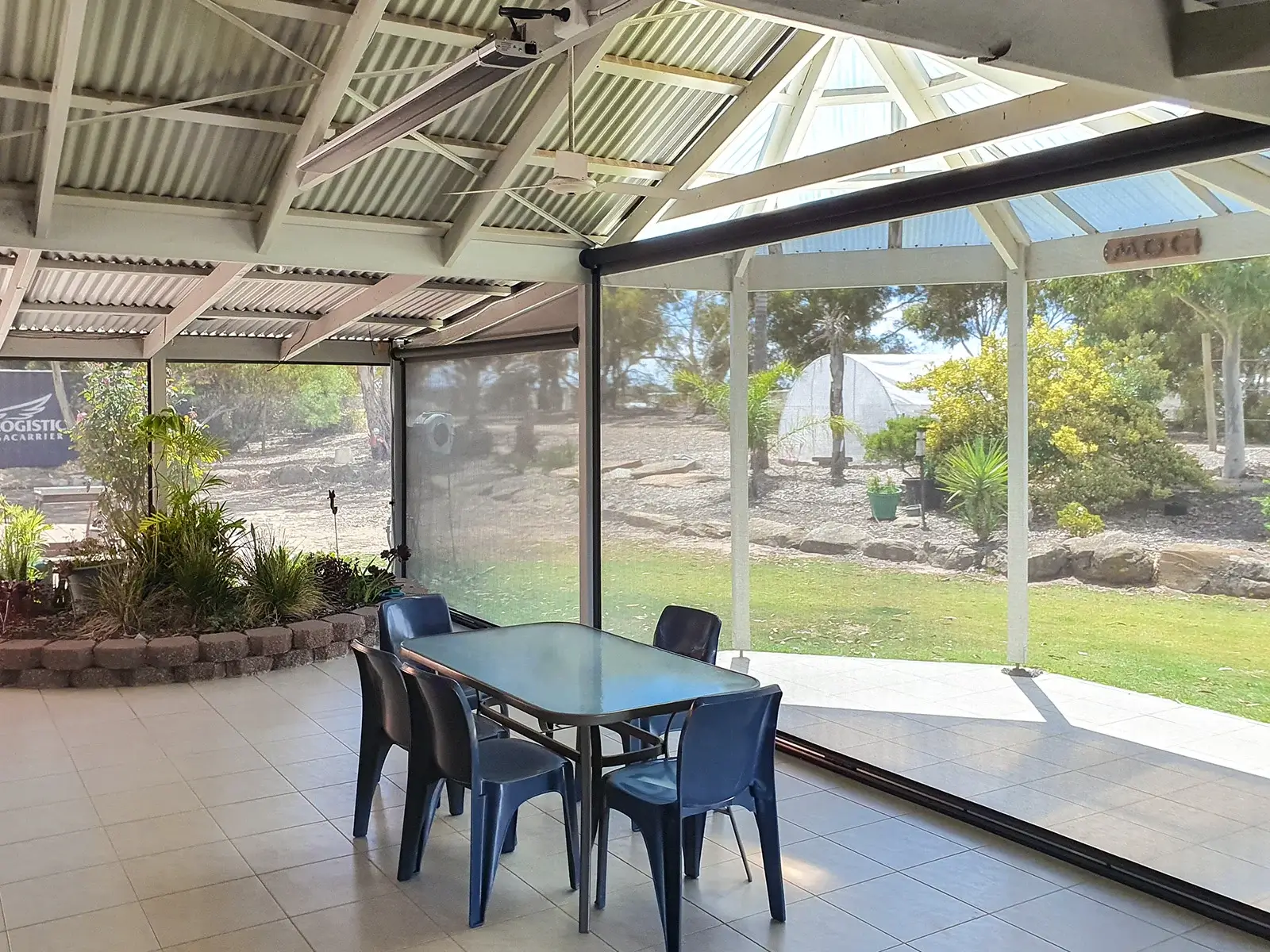 Spacious outdoor patio with outdoor blinds, featuring a dining table with blue chairs, surrounded by garden plants and offering a view of a landscaped yard.