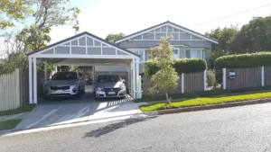 House with a neatly trimmed hedge and fence, featuring a timber carport over the driveway with two parked cars.