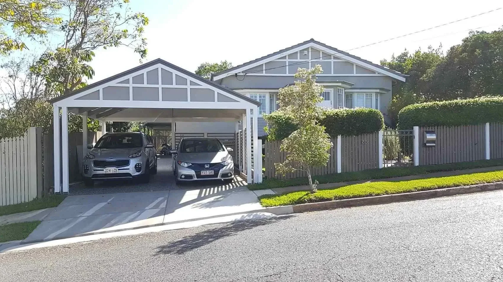 House with a neatly trimmed hedge and fence, featuring a timber carport over the driveway with two parked cars.