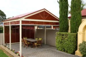 Outdoor dining area under a timber pergola with a gabled roof and horizontal slats, featuring a wooden table and chairs on a paved patio, surrounded by tall trees and greenery.